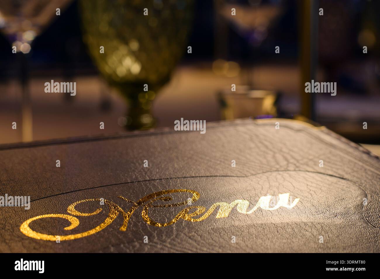 Menu rilegato in pelle con lettere dorate lucide al tavolo del ristorante. Raffinata atmosfera da pranzo con tavolo, bicchieri defocalizzati, atmosfera accogliente Foto Stock