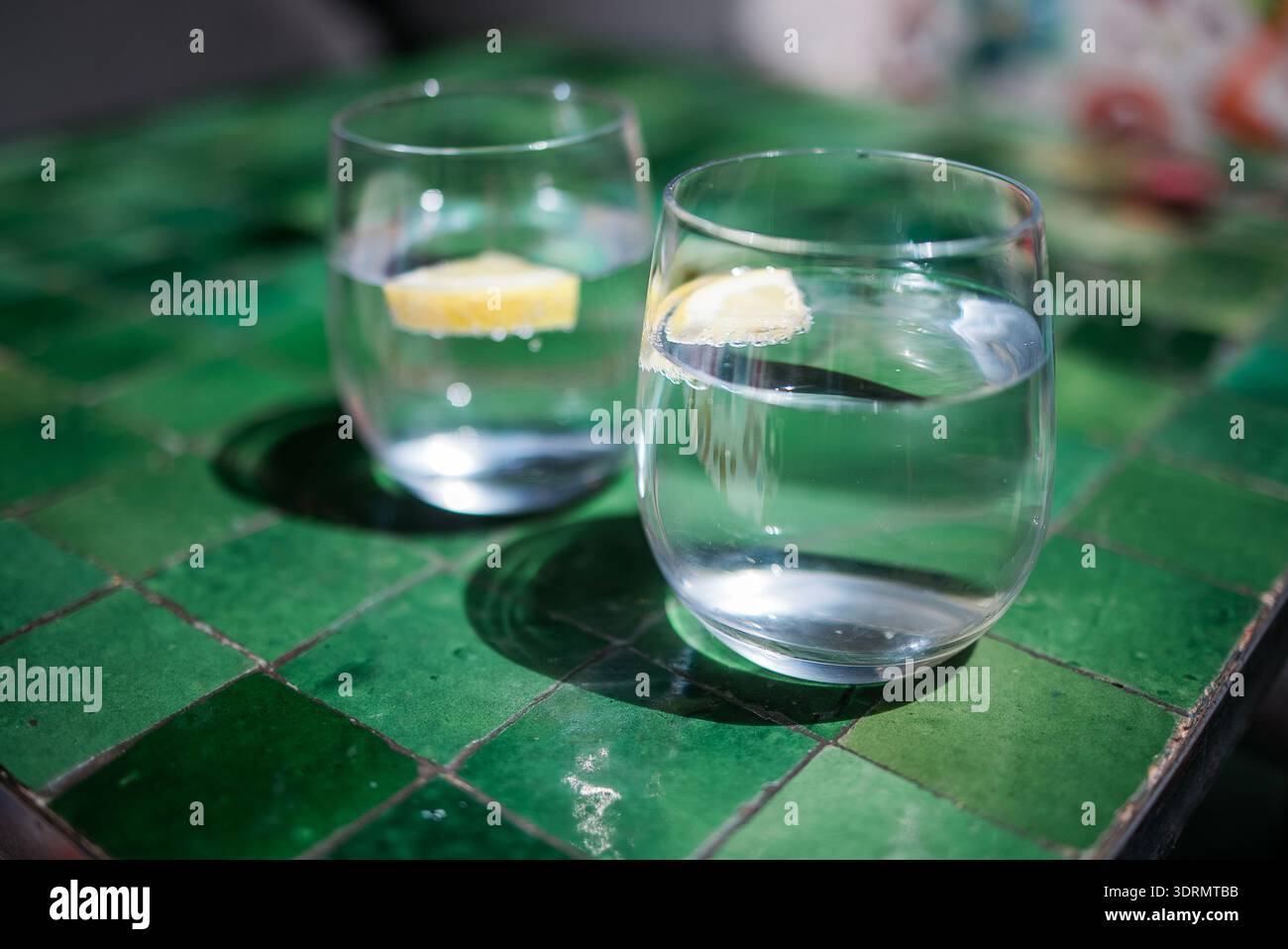 Due bicchieri di acqua limpida con fette di limone si trovano su un vivace tavolo piastrellato verde, che offre bevande rinfrescanti in un luminoso e. Foto Stock
