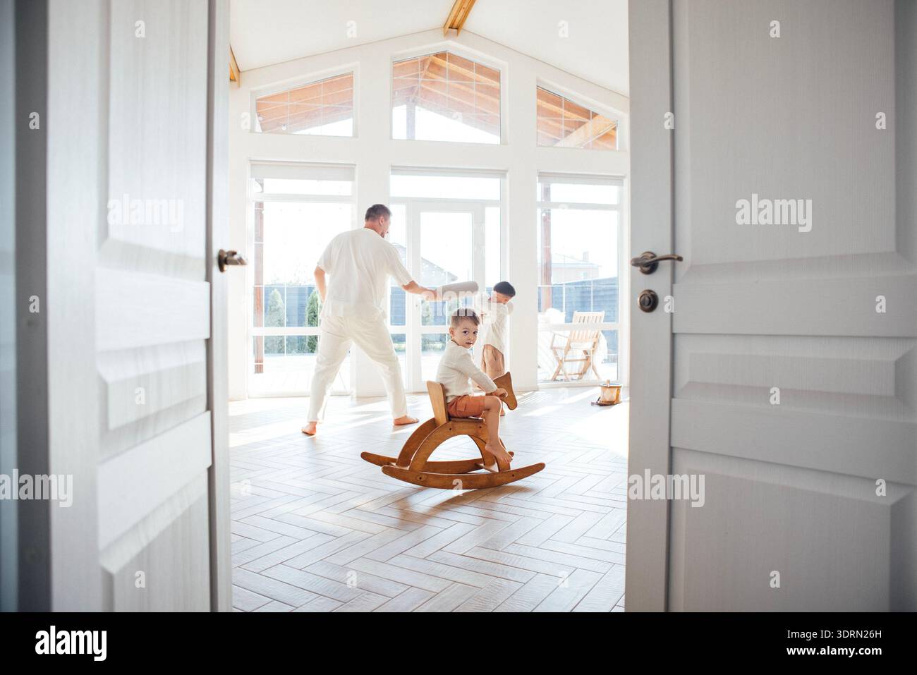 Bambini a dondolo in legno in una stanza luminosa, mentre gli adulti giocano e ballano, creando un'atmosfera gioiosa di legame familiare e di happ Foto Stock