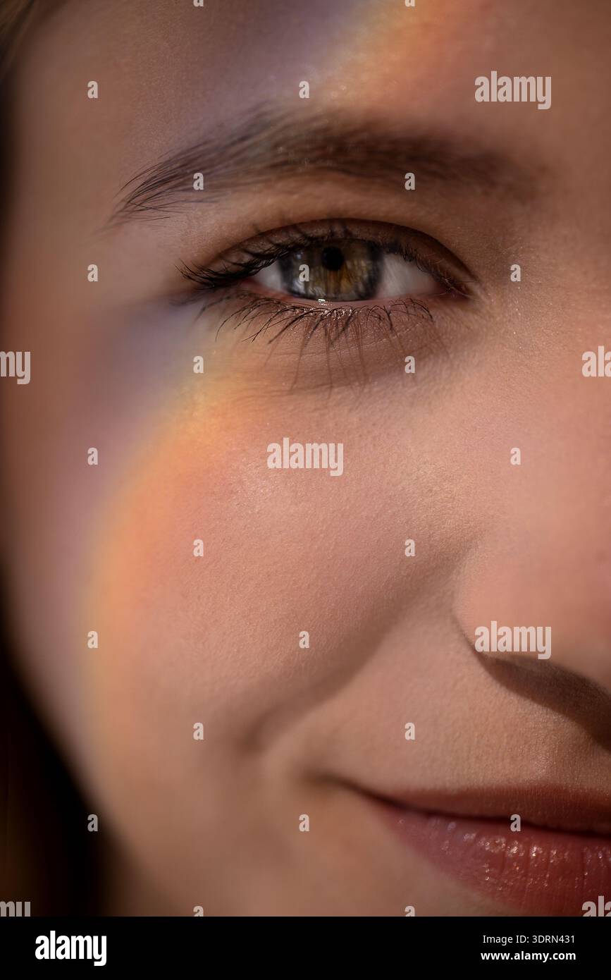 Ritratto ravvicinato di una giovane donna con un sottile riflesso arcobaleno sulla guancia, che mostra i suoi occhi luminosi e un sorriso morbido, cappello Foto Stock