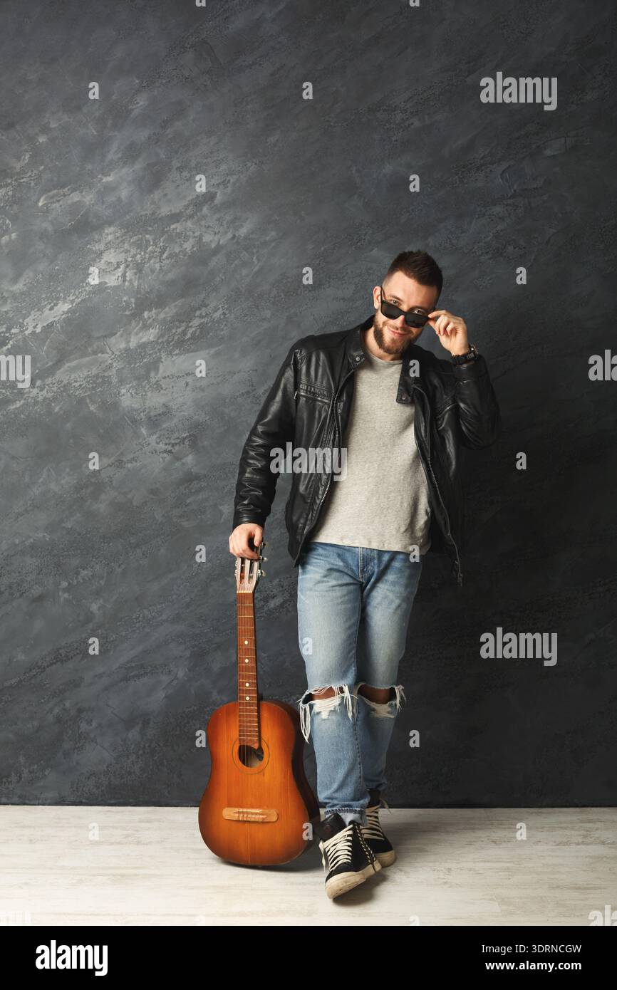 L'uomo è in piedi con la chitarra contro una parete scura, indossa occhiali da sole e una giacca in pelle Foto Stock