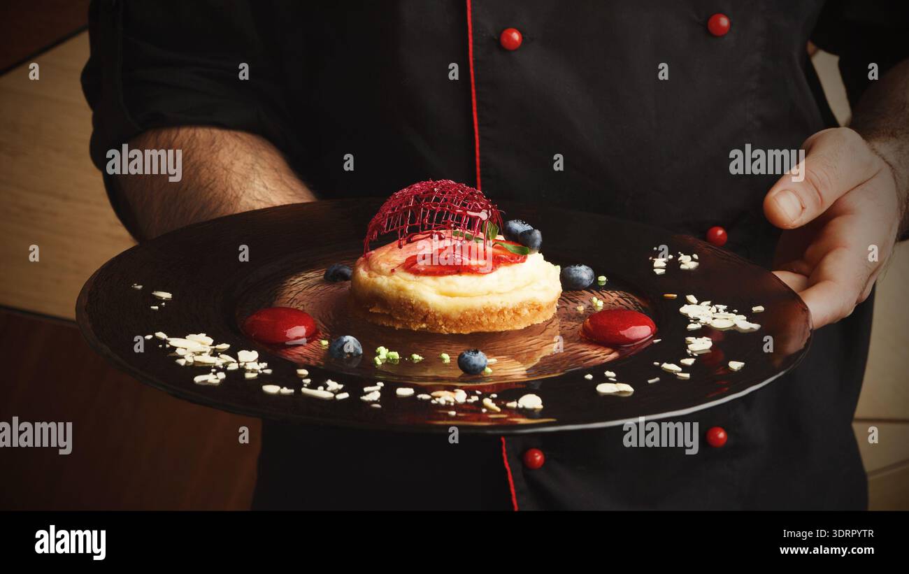 Lo chef presenta dessert su piatto con frutti di bosco e salsa in un ambiente culinario Foto Stock