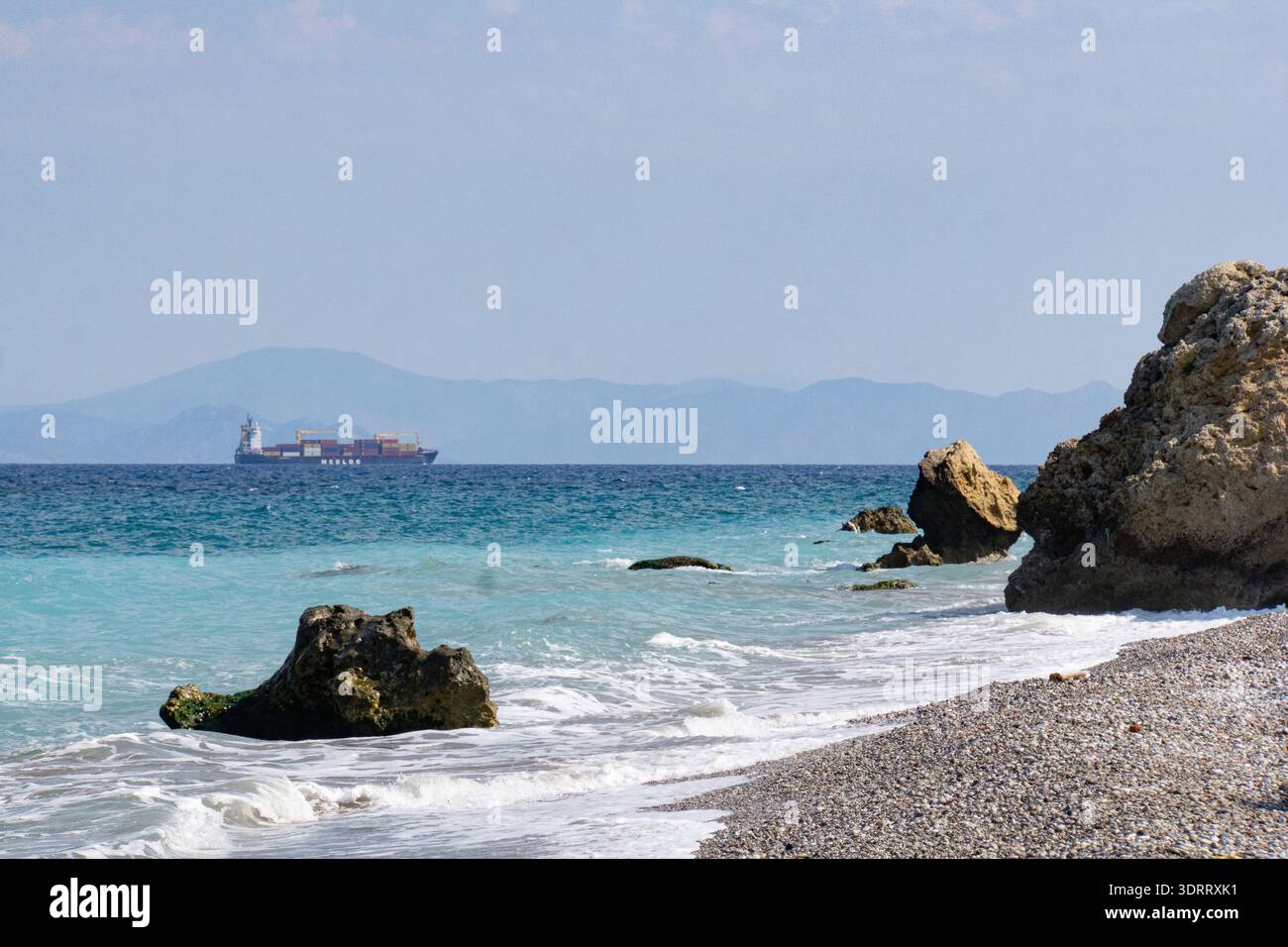 Costa rocciosa sull'isola di Rodi, con mare turchese, onde dolci e una nave da carico che naviga all'orizzonte sotto un cielo limpido. Foto Stock