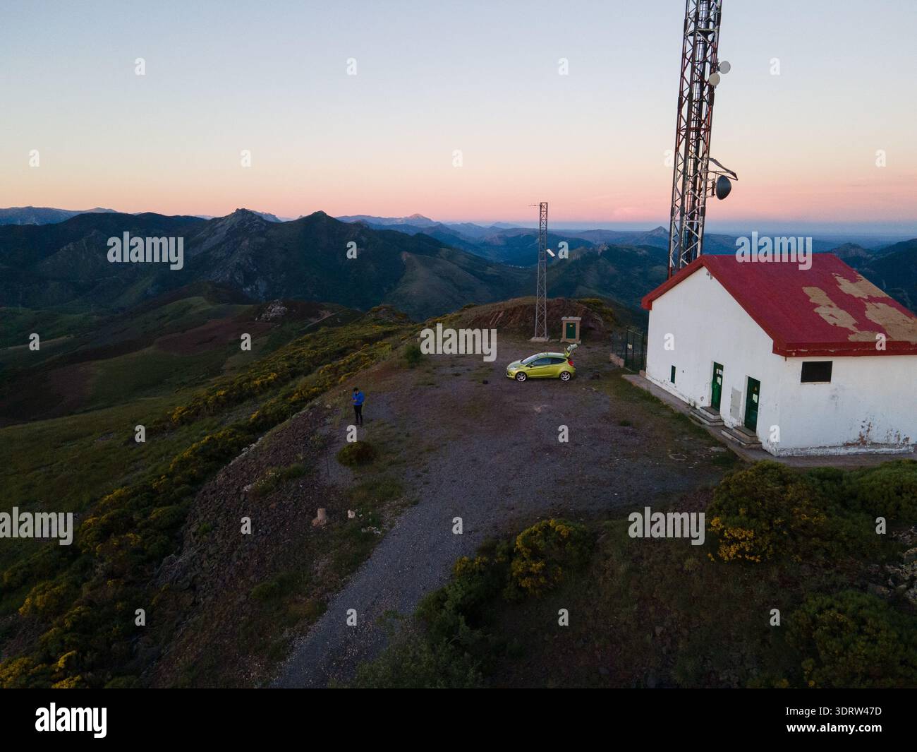 Vista aerea di una figura solitaria e di un'auto gialla vicino a un edificio bianco con un tetto rosso in cima a una montagna, in contrasto con le sfumature morbide del cielo del tramonto, Foto Stock