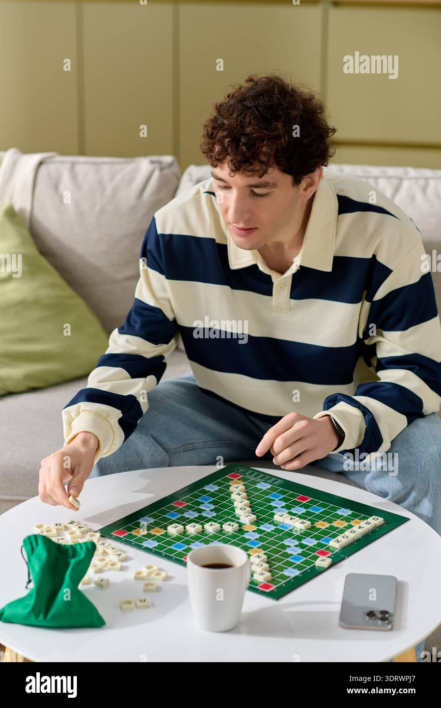 Un bel giovane si concentra intensamente sul suo gioco di Scrabble a casa, circondato da un'atmosfera accogliente. Foto Stock