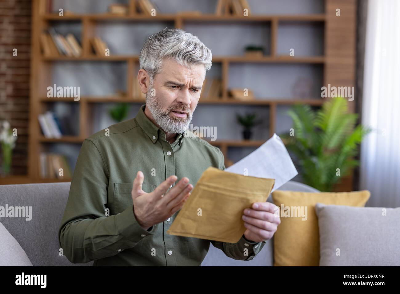 Uomo dai capelli grigi maturo seduto su un divano, tenendo in mano una busta aperta con un'espressione preoccupata e frustrata dopo aver letto a casa la posta economica o ufficiale Foto Stock