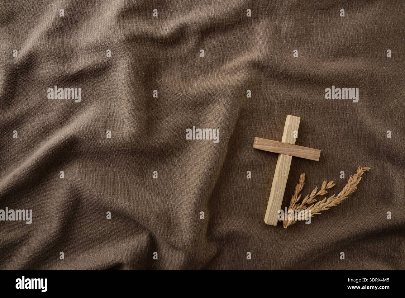 Una croce di legno posta su un tessuto marrone rugoso con steli di grano, simboleggia la fede cristiana, la semplicità e la raccolta in un ambiente rustico e caldo. Foto Stock