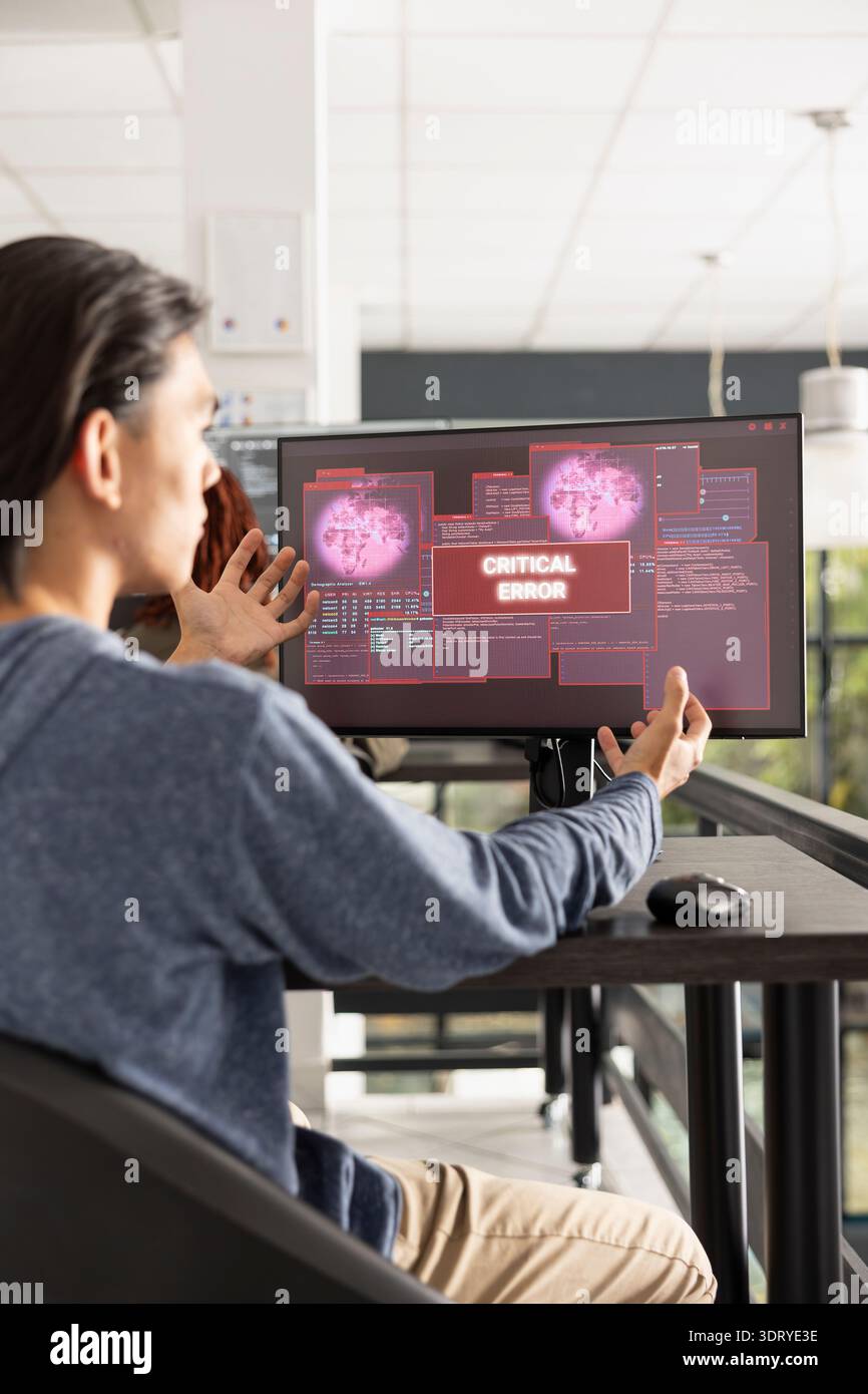Ritratto del programmatore maschio che gesta in caso di allarme di errore sul monitor desktop. Lo sviluppatore di software asiatico si trova alla scrivania in un ufficio moderno, reagendo ai messaggi di errore critici sullo schermo del computer. Foto Stock