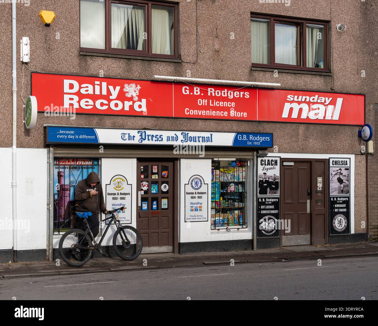 Una persona con una giacca marrone con cappuccio si stende con una bicicletta nera, guardando un telefono cellulare. Ciò si verifica al di fuori di G.B. Rodgers Off Licence Convenience sto Foto Stock