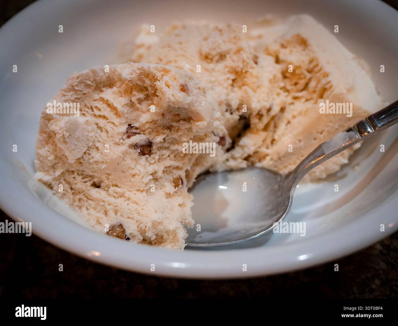 Primo piano del gelato al burro pecan in un recipiente bianco con un cucchiaio, mostrando una consistenza cremosa e pezzi di noci Foto Stock
