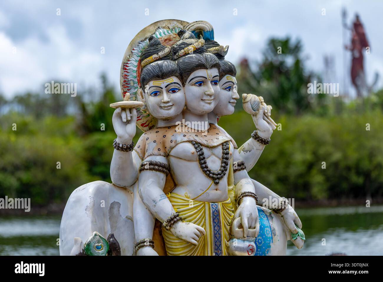 Dettagliata statua di Trimurti con facce e braccia multiple, adagiata su una vegetazione lussureggiante Foto Stock