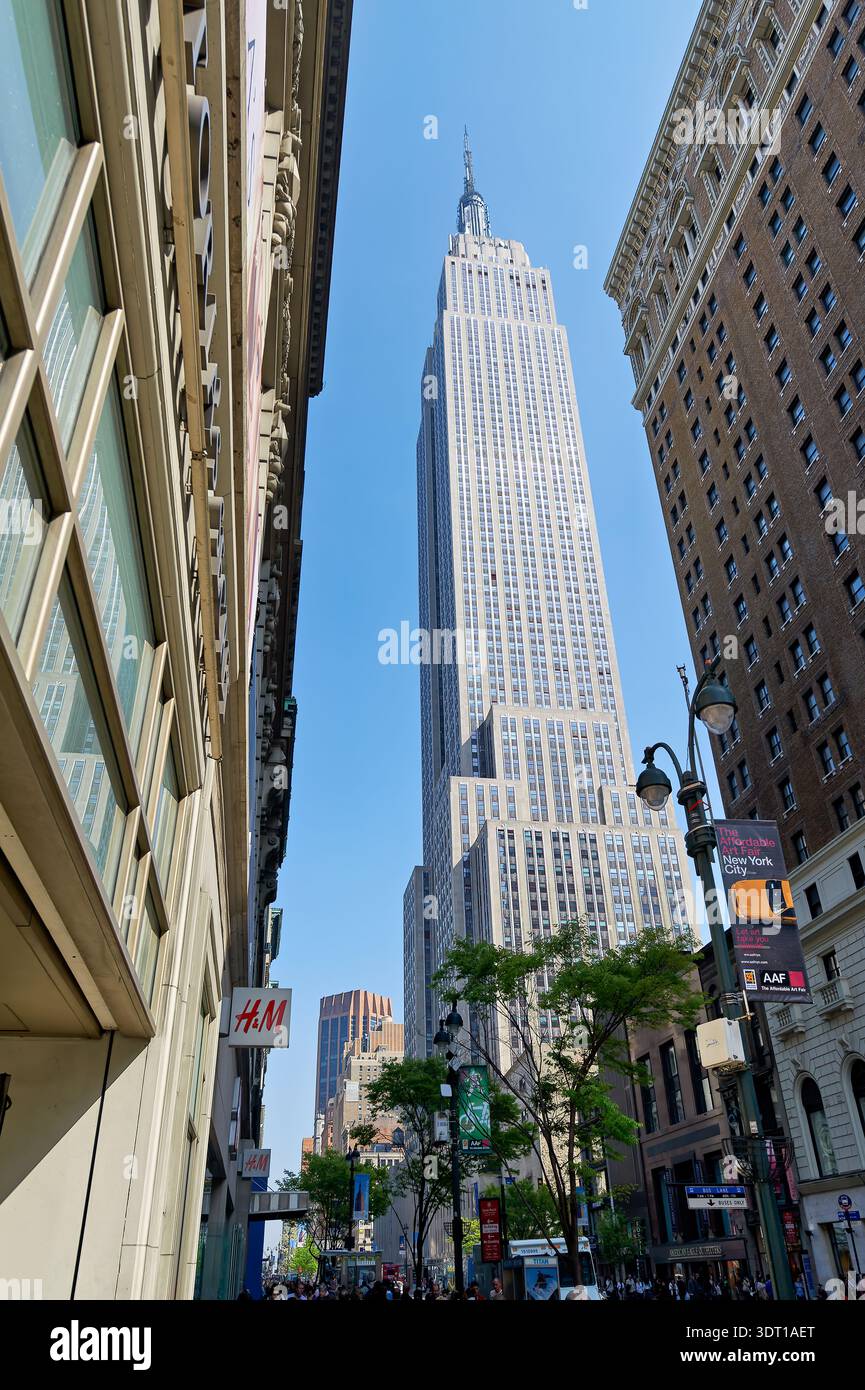 Le persone passeggiano a Manhattan mentre l'Empire State Building si erge sopra di loro sotto un cielo azzurro. Molti negozi ed edifici circondano l'area. Foto Stock