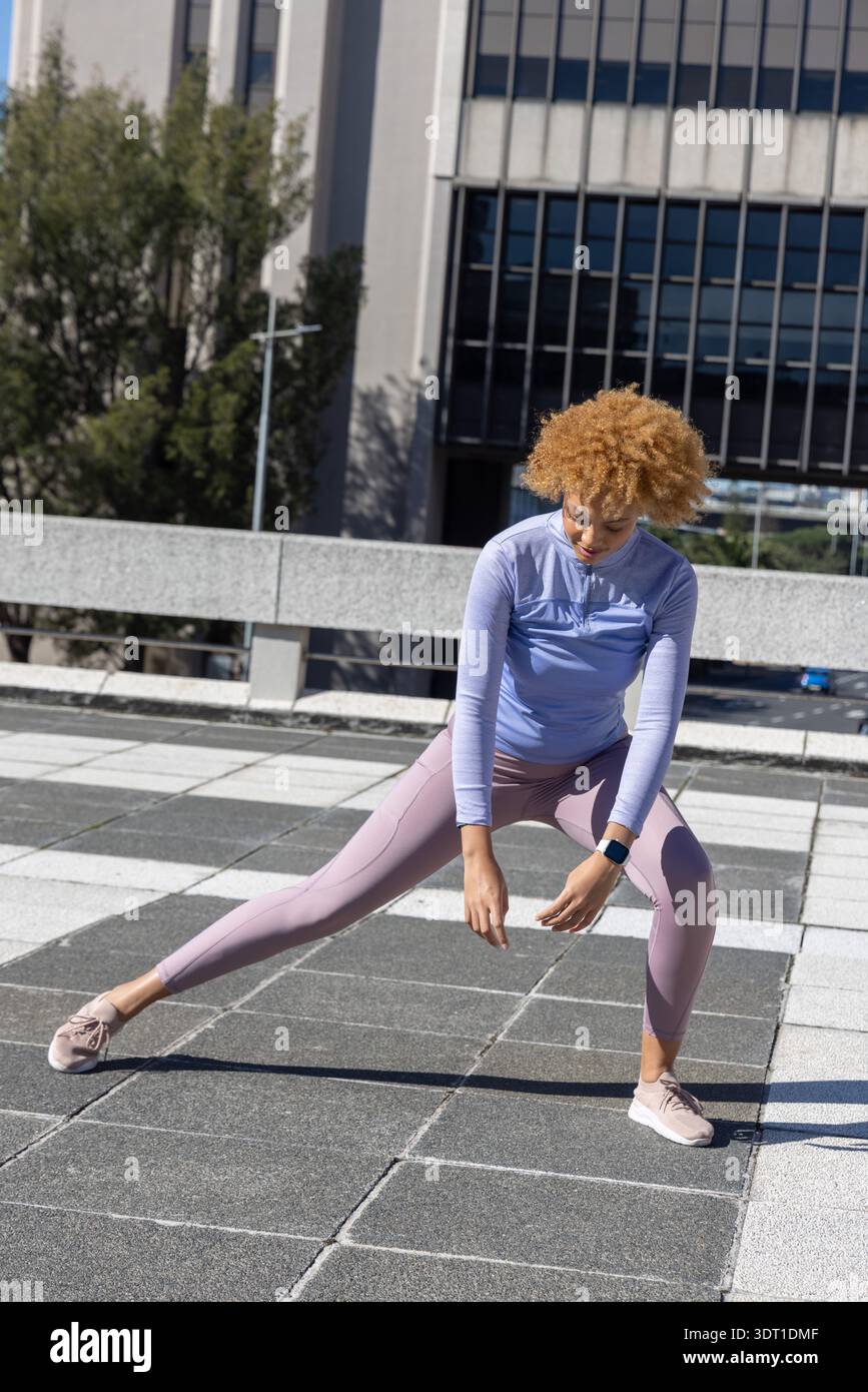 Donna afroamericana che fa l'affondo laterale sul tetto in tegole indossando leggings e smartwatch Foto Stock