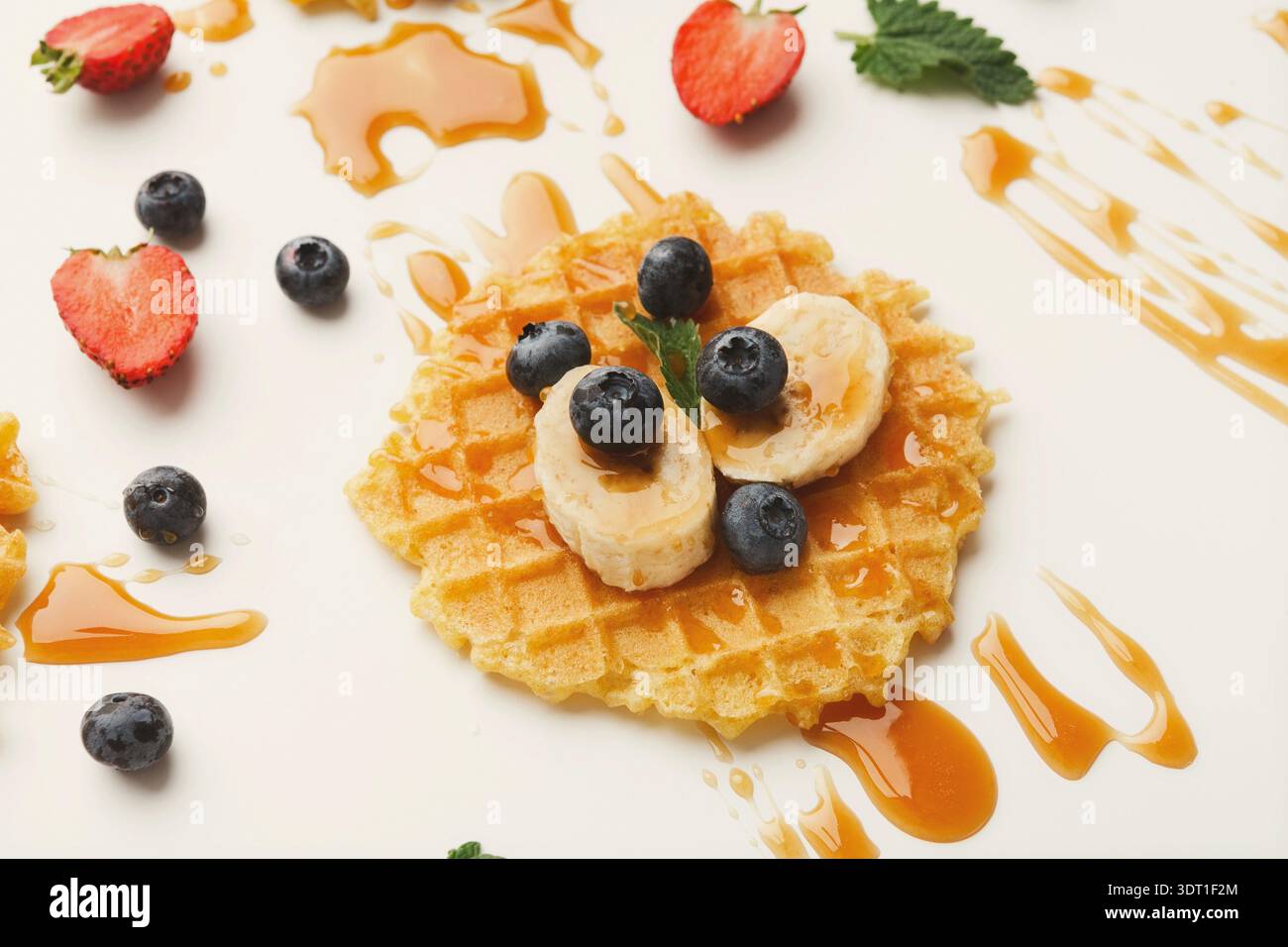 Waffle con banana e frutti di bosco su sfondo bianco con sciroppo sparso intorno Foto Stock