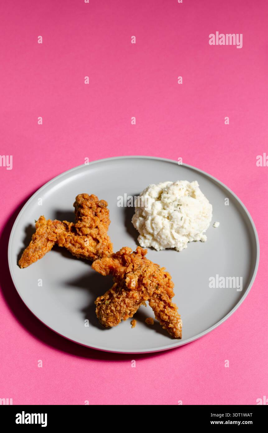 Una deliziosa porzione di ali di pollo croccanti abbinate a cremosi purè di patate è esposta su un piatto grigio su un vivace sfondo rosa, la creatina Foto Stock