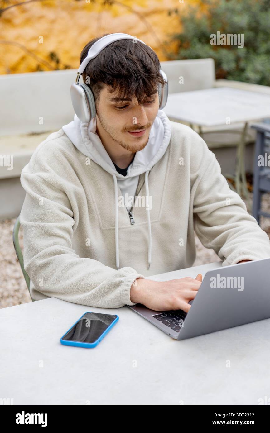 Un giovane uomo siede all'aperto, indossa le cuffie e lavora su un notebook. Accanto a lui c'è uno smartphone sul tavolo. Questa scena cattura l'essenza del mod Foto Stock