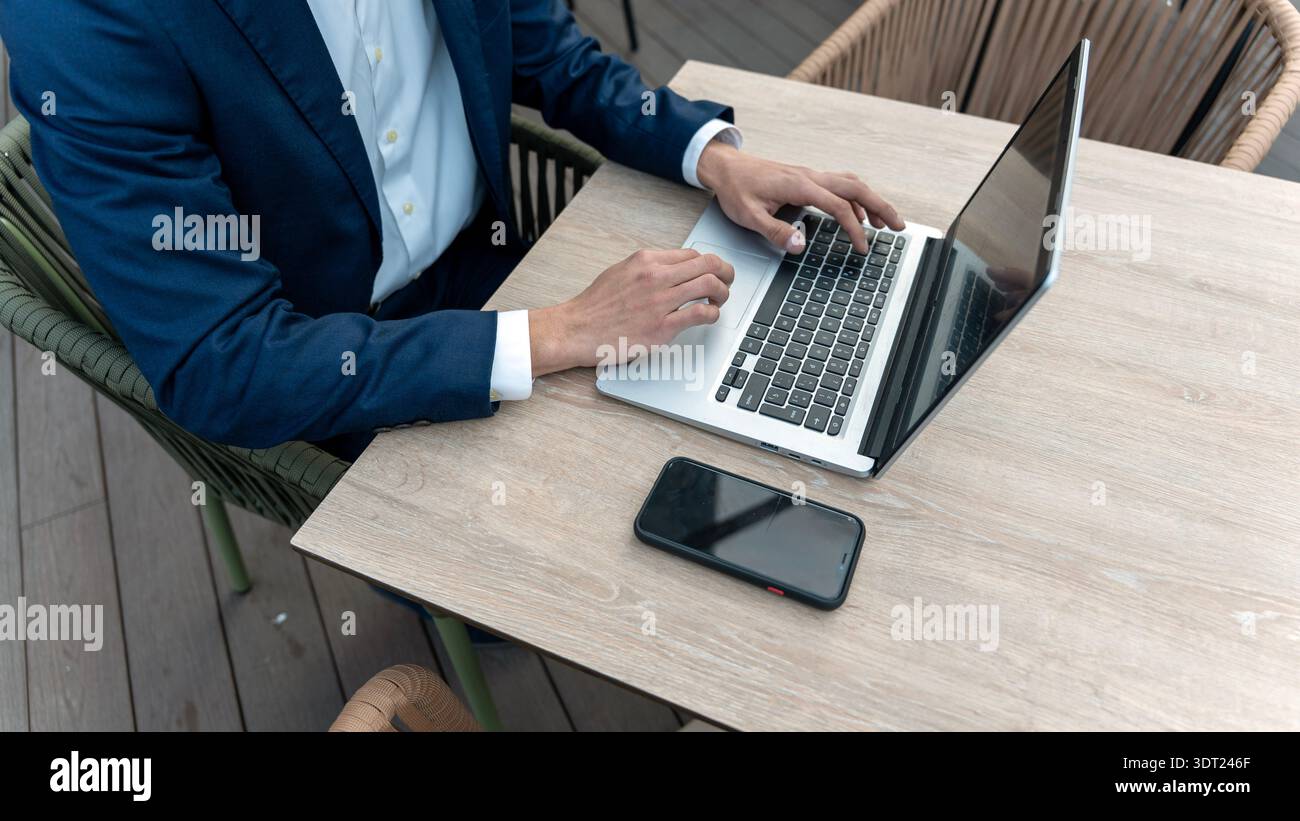 Uomo d'affari in tuta blu che lavora su un computer portatile su un tavolo di legno, con un telefono cellulare accanto a lui. L'ambiente è un moderno spazio di lavoro all'aperto, focalizzato Foto Stock