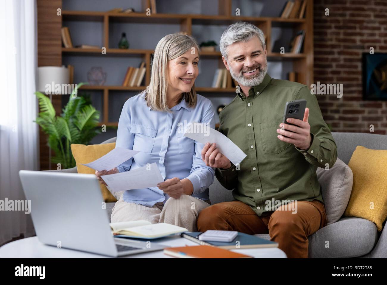 Una coppia matura sorridente che gestisce le finanze personali e le bollette insieme a casa, guardando uno schermo di uno smartphone con documenti cartacei, un computer portatile e una calcolatrice sul tavolino da caffè Foto Stock