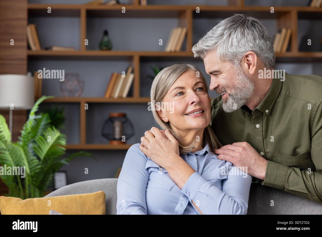 Il marito senior abbraccia delicatamente la moglie sorridente nel loro accogliente soggiorno, condividendo un momento caldo e affettuoso che riflette amore duraturo, fiducia e compagnia confortevole Foto Stock