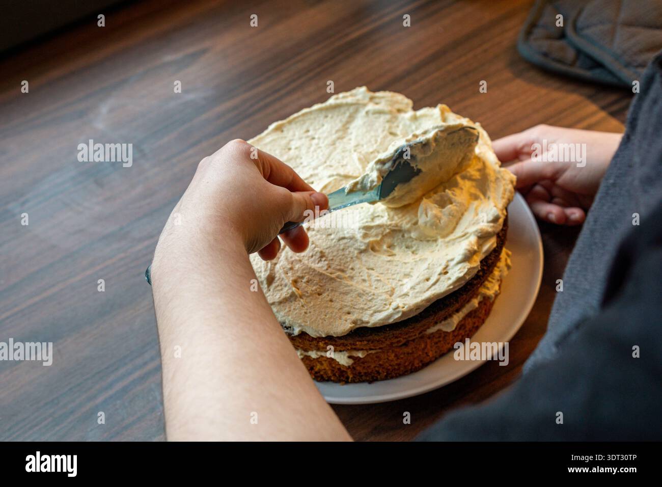 Vista laterale di una persona che usa una spatola per lisciare la panna su una torta fatta in casa. Foto Stock
