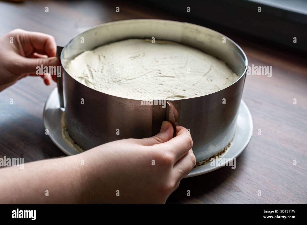Vista ravvicinata delle mani che fissano un anello regolabile in metallo intorno a una torta a strati appena smerigliata. Foto Stock