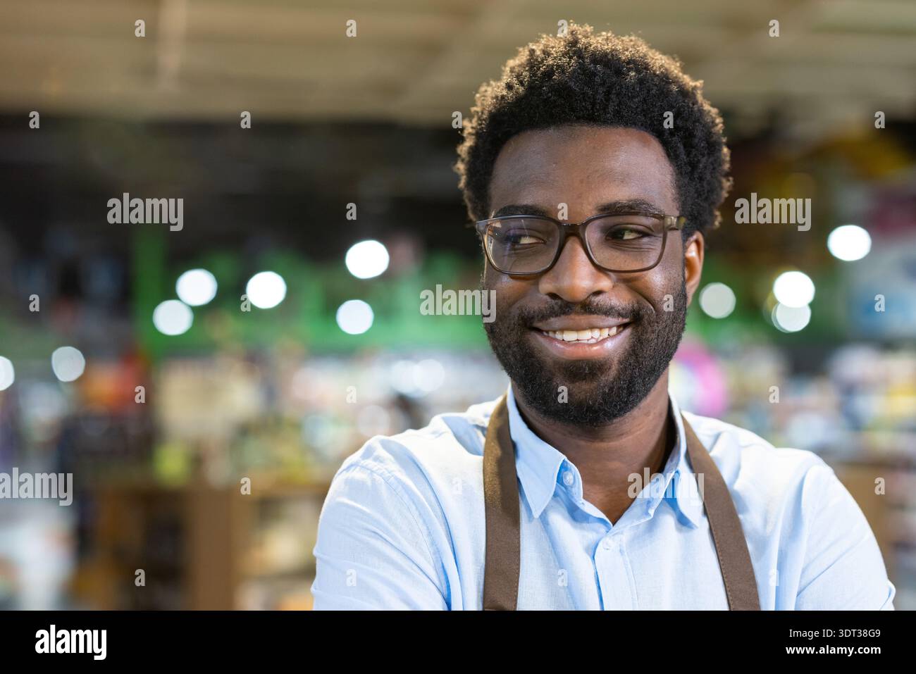 Uomo allegro che indossa occhiali e grembiule, sorride e guarda lontano mentre si trova nel suo negozio di piccole imprese, rappresentando il successo e l'imprenditorialità Foto Stock