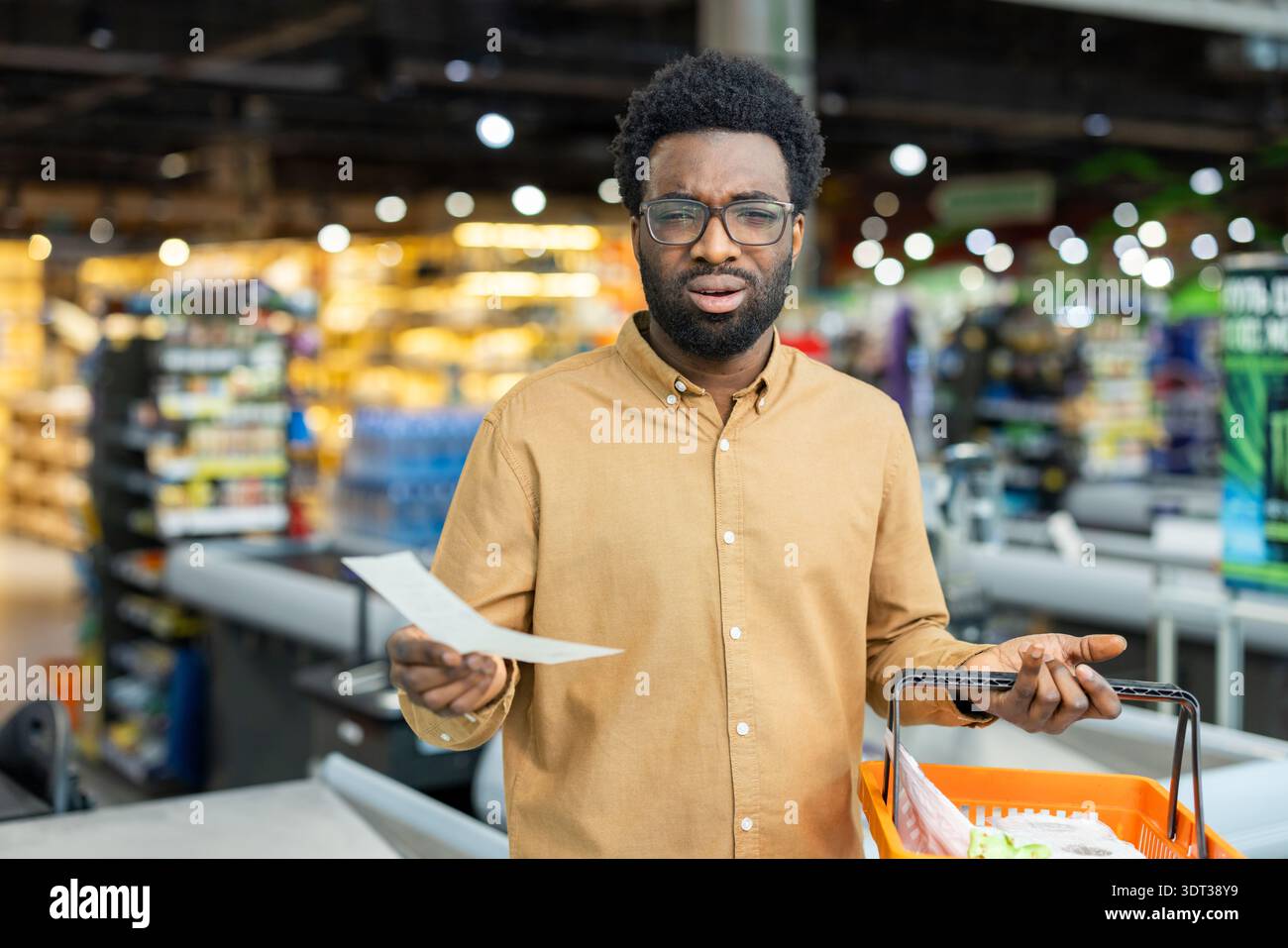 Uomo afroamericano alla cassa del supermercato con una lunga ricevuta e un piccolo paniere, confuso e frustrato dagli alti prezzi e dall'aumento dell'inflazione che influenzano gli acquisti quotidiani Foto Stock