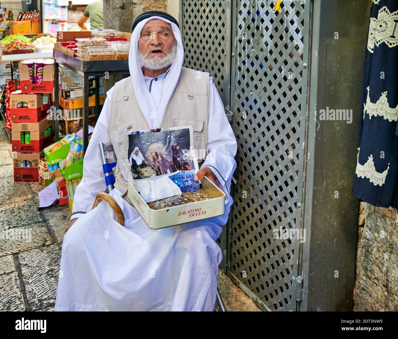 Nella città vecchia di Gerusalemme, accanto a un negozio si trova un mendicante. Tiene in mano un vassoio con vari articoli in vendita. La gente passa senza fermarsi. Foto Stock