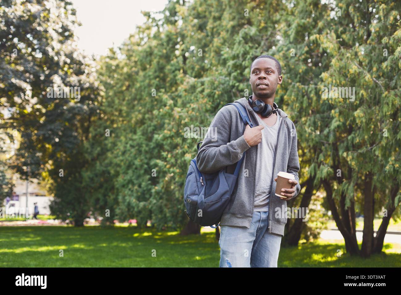 Young Man si trova a Park e tiene il caffè, indossa cuffie e zaino in una giornata di sole Foto Stock
