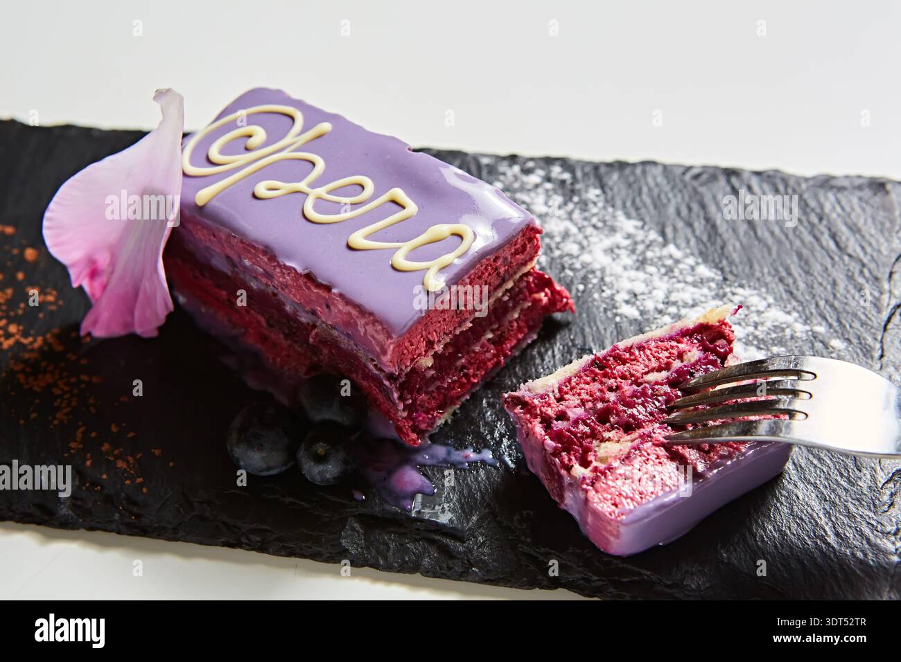 Moderna fetta di torta Opera con vibrante glassa viola, delicata calligrafia al cioccolato bianco e mirtilli freschi, un dessert multi-sensoriale che mette in risalto Foto Stock