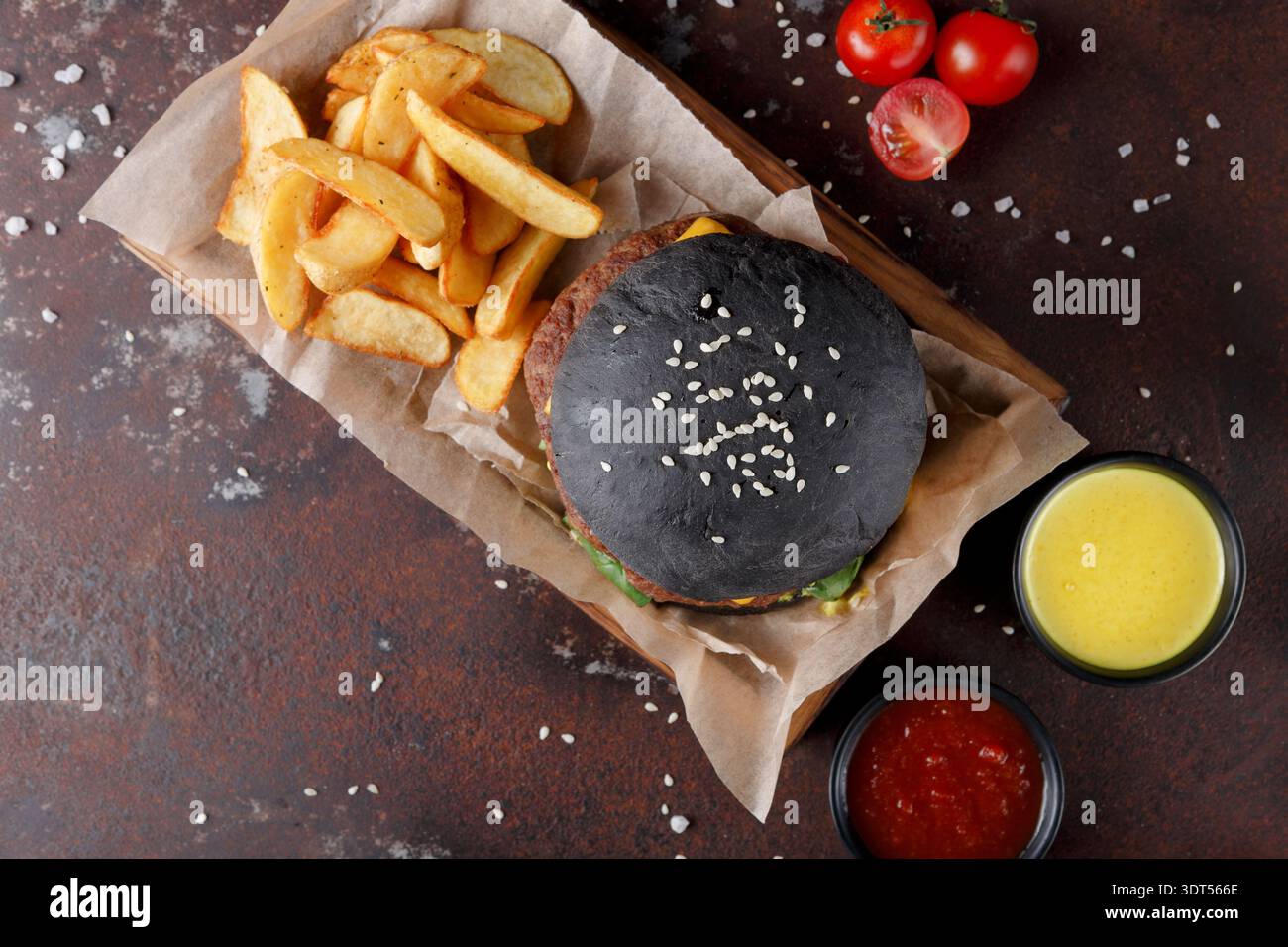Delizioso hamburger con panino nero, patatine fritte e salse servito su vassoio di legno con pomodori freschi Foto Stock