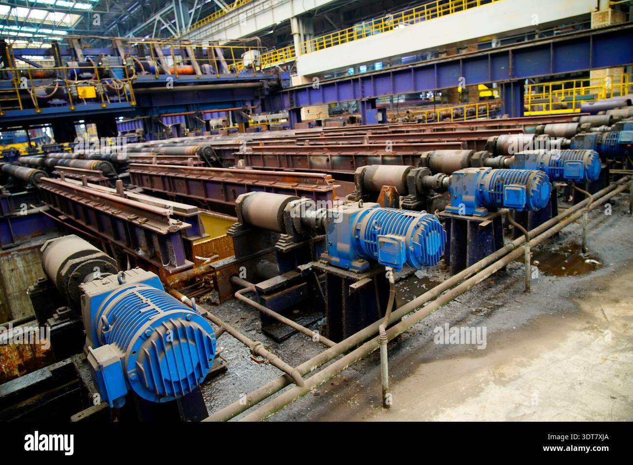 L'area di produzione principale di laminatoio a caldo, l'array di motori di azionamento blu e la tavola dei rulli di laminazione arrugginiti costituiscono l'officina industriale di formalina in acciaio Foto Stock