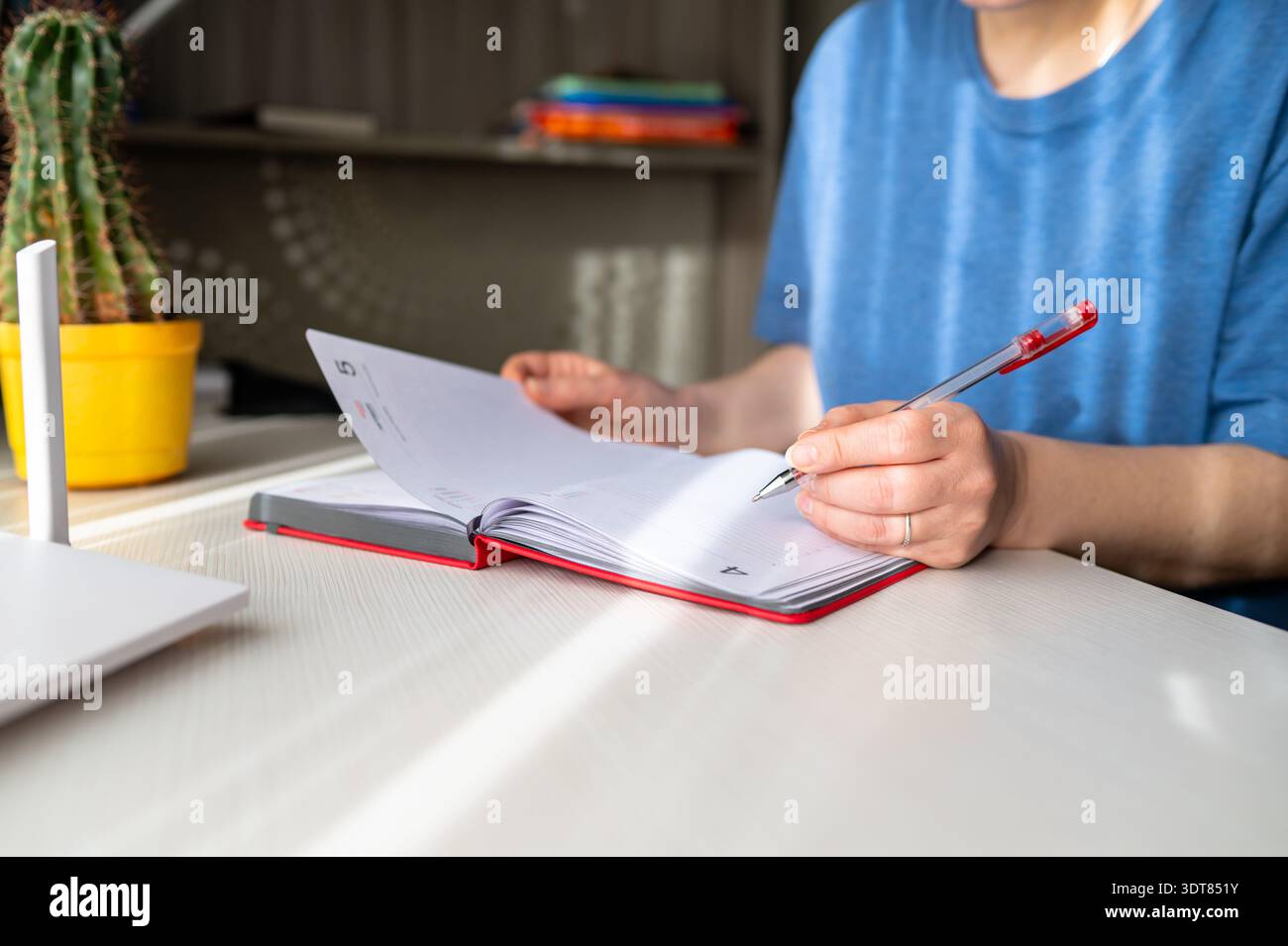 Primo piano di una donna che scrive a mano in un personal planner rosso con una penna rossa su una scrivania bianca. Concetto di ufficio domestico, pianificazione, journaling e WO remoto Foto Stock