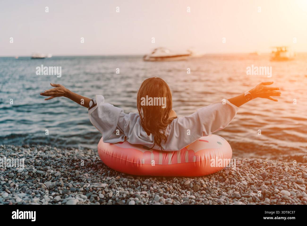 Donna spiaggia oceano. Donna rilassante seduta sulla spiaggia di ciottoli, anello gonfiabile, che abbraccia la vasta vista dell'oceano all'alba. Foto Stock