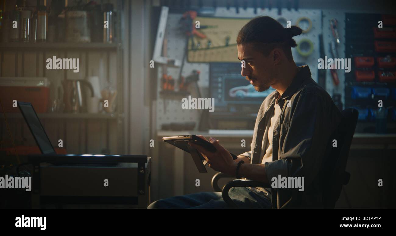 Specialista automobilistico maschio seduto in officina, che utilizza la configurazione a doppio schermo con laptop per fare riferimento incrociato ai dati del motore durante l'ispezione approfondita del veicolo. Concetto di soluzioni diagnostiche intelligenti. Foto Stock