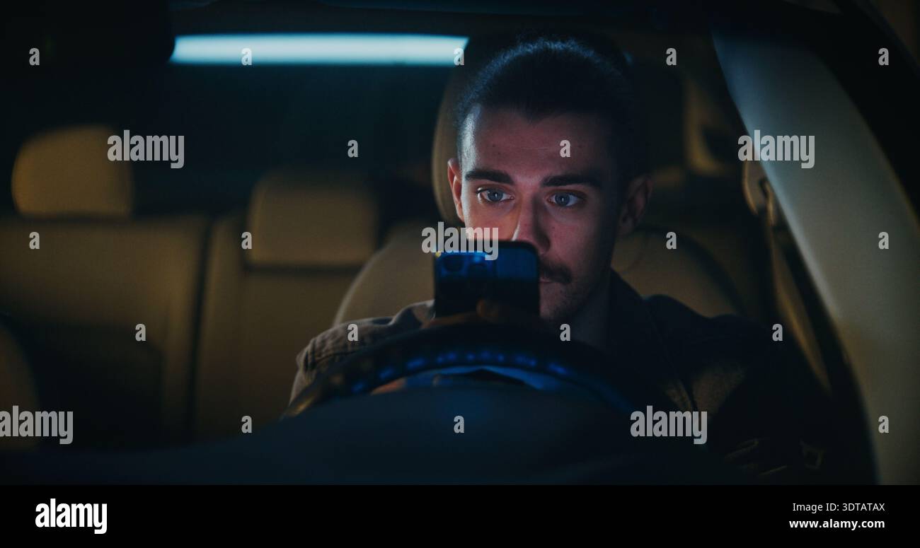 Un ragazzo con capelli lunghi legati alla schiena siede in auto, leggendo attentamente i messaggi sullo smartphone. L'uomo fa una pausa ponderata prima di digitare la risposta e si impegna in una conversazione importante e significativa. Comunicazione sociale. Foto Stock