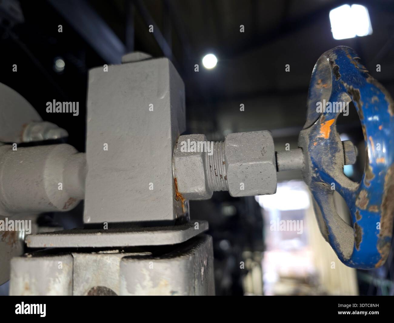 Un primo piano mostra una valvola collegata a un raccordo per tubi all'interno di uno spazio industriale buio. Le superfici metalliche sono usurate e la luce evidenzia i dettagli. Foto Stock