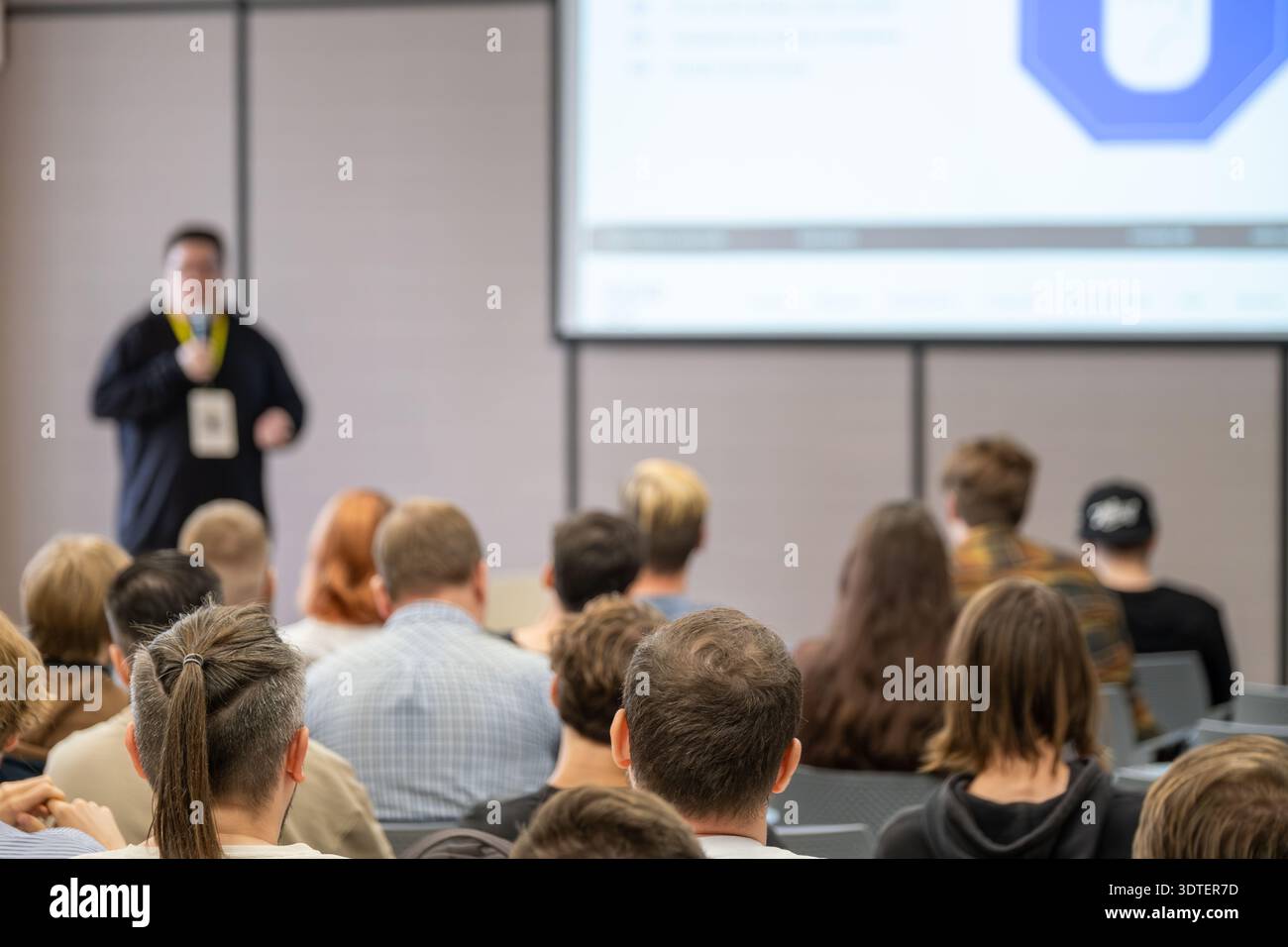 Il pubblico ascolta il relatore che presenta nella moderna sala conferenze. Il grande schermo del proiettore visualizza le diapositive mentre i partecipanti si concentrano sulla presentazione. Foto Stock