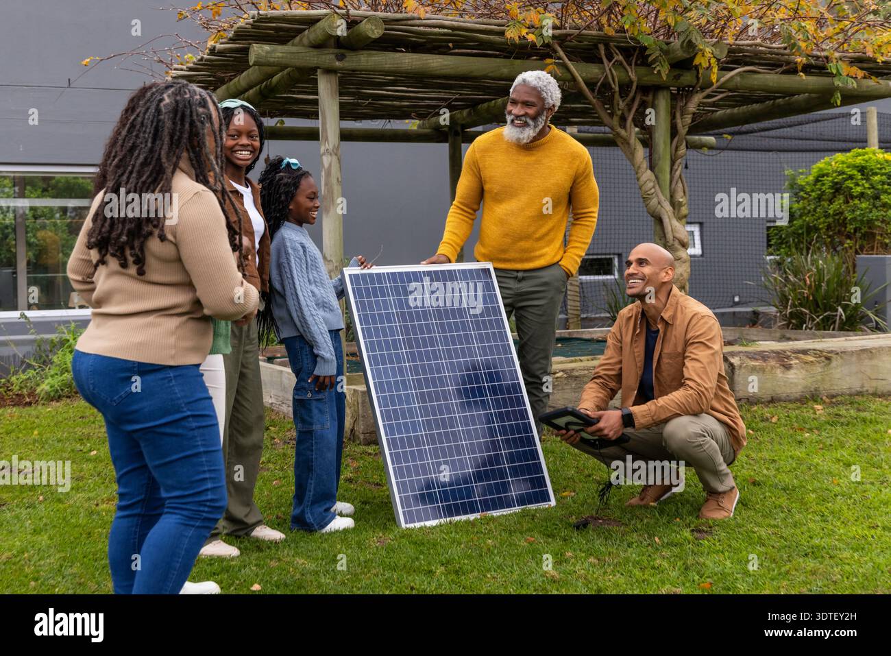 Famiglia afroamericana che prova il pannello solare sul prato, senior steadying, inginocchiato che tiene la tavoletta Foto Stock