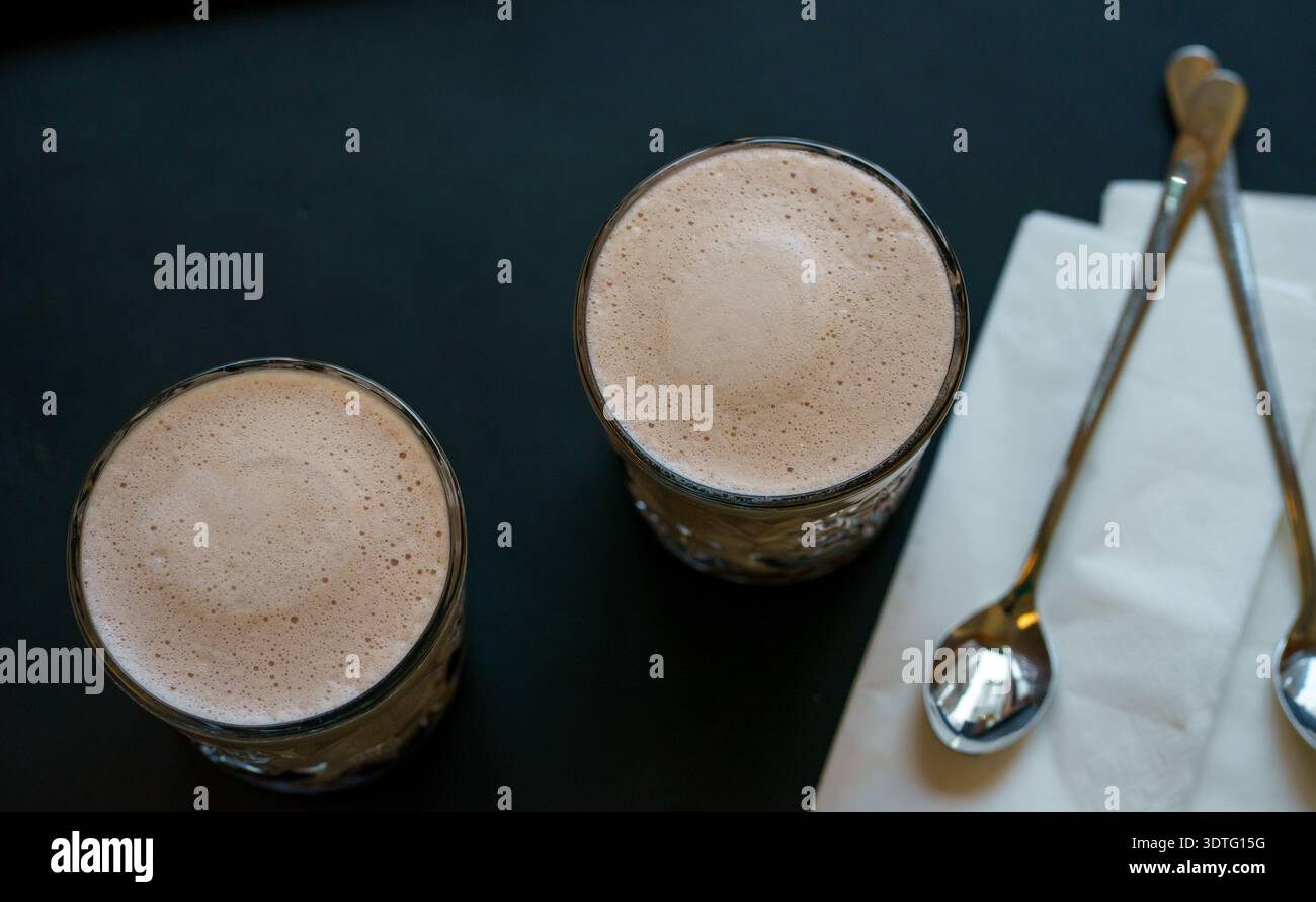 Vista dall'alto di due bicchieri di cremosa cioccolata calda con schiuma di latte liscia, serviti con cucchiai e tovagliolo. Accogliente atmosfera da caffetteria e concetto di bevanda calda. Foto Stock