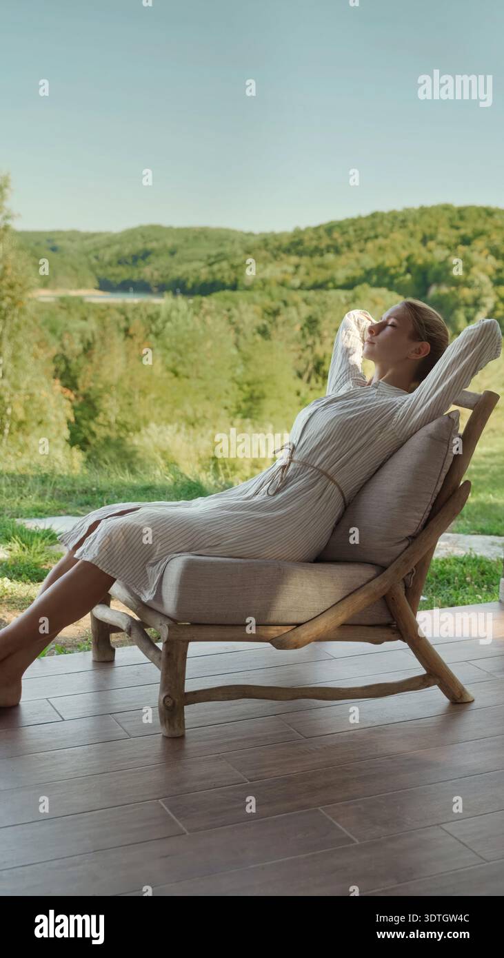 Una donna con un abito bianco si appoggia su una sedia di legno, godendosi la tranquillità di un lussureggiante paesaggio verde in una giornata di sole. Foto Stock