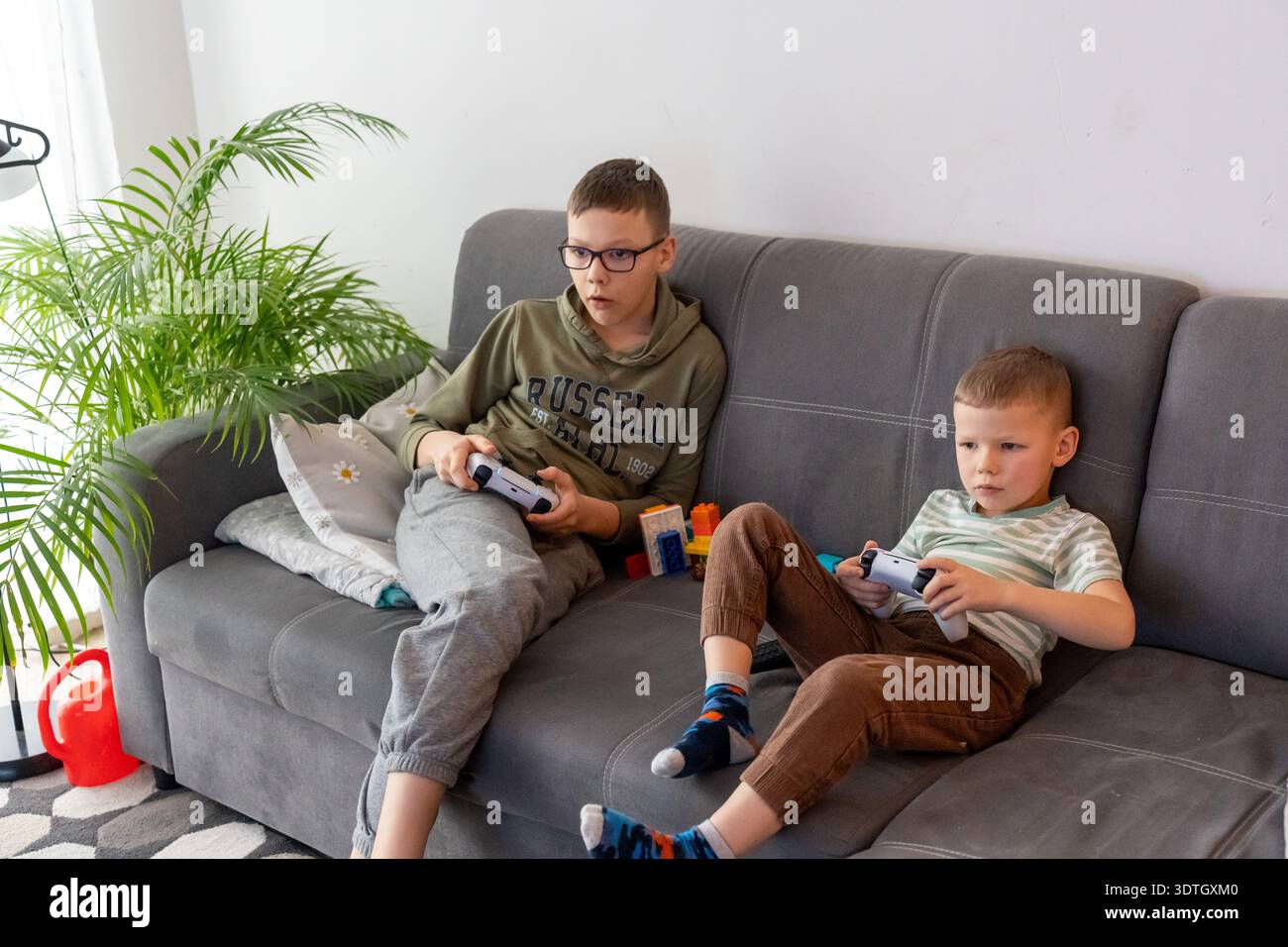 Due fratelli concentrati in abiti casual che giocano ai videogiochi con controller wireless su un divano grigio con giocattoli colorati. Concetto di rivalità tra fratelli, vide Foto Stock