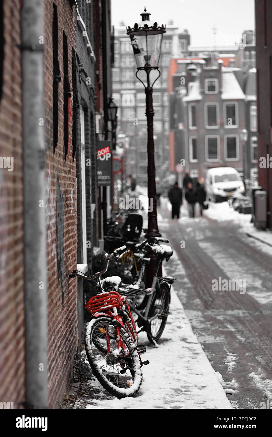 La neve si aggrappa alle biciclette in una stretta strada di Amsterdam, Paesi Bassi, mentre una bici rossa si distingue vicino alle pareti di mattoni e i pedoni camminano in lontananza. Foto Stock