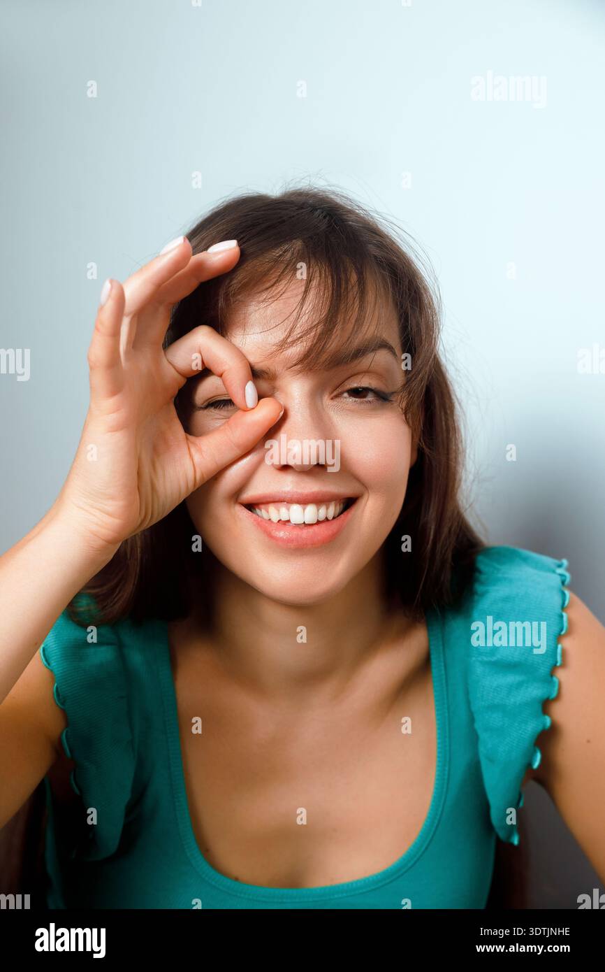 Una donna siede con un'espressione giocosa sul viso, usando la mano per formare un cerchio su un occhio mentre sorride. Ha i capelli lunghi e indossa una t semplice Foto Stock