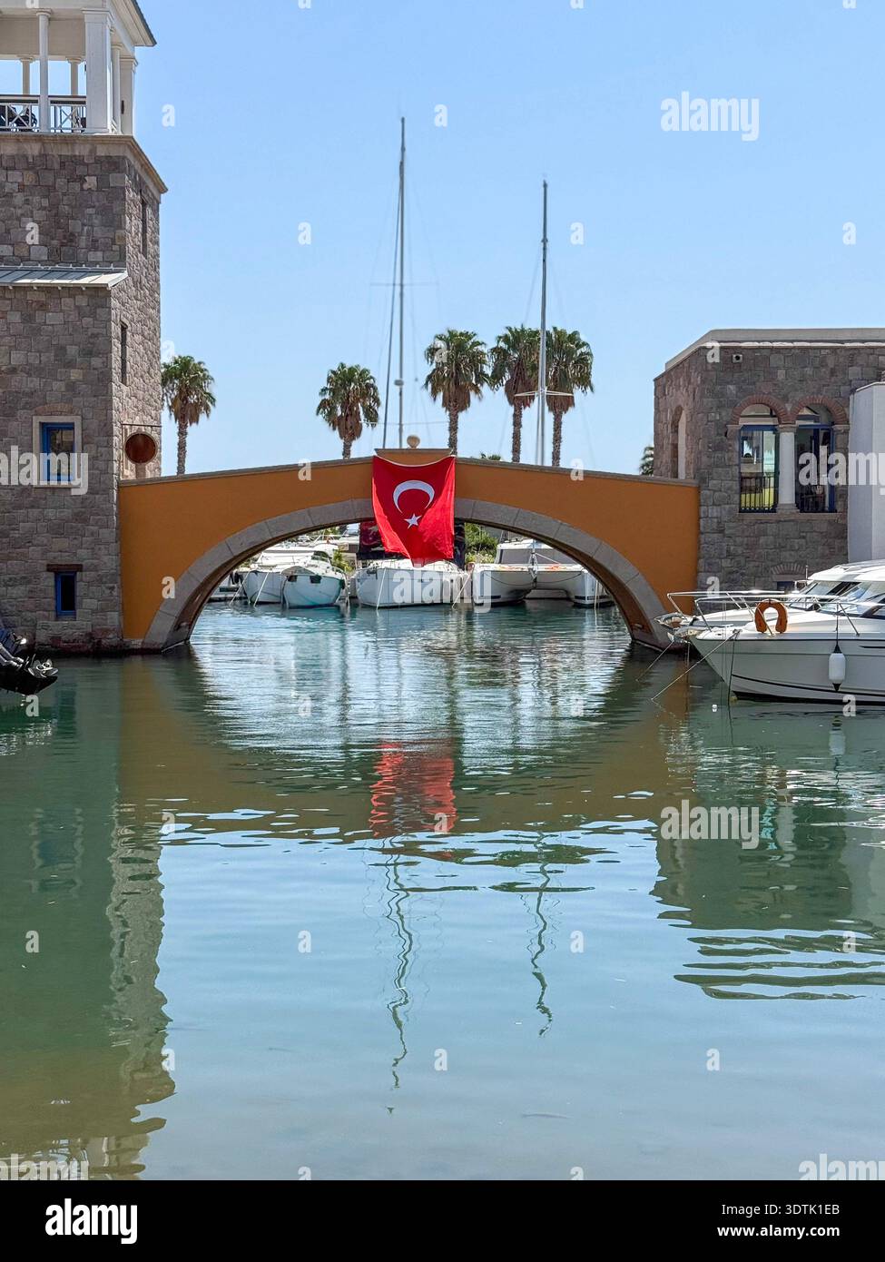 Un tranquillo porticciolo con un ponte ad arco di pietra drappeggiato da una bandiera turca. Palme estive e barche a vela completano l'idilliaco scenario delle vacanze sotto un chiaro bagno Foto Stock