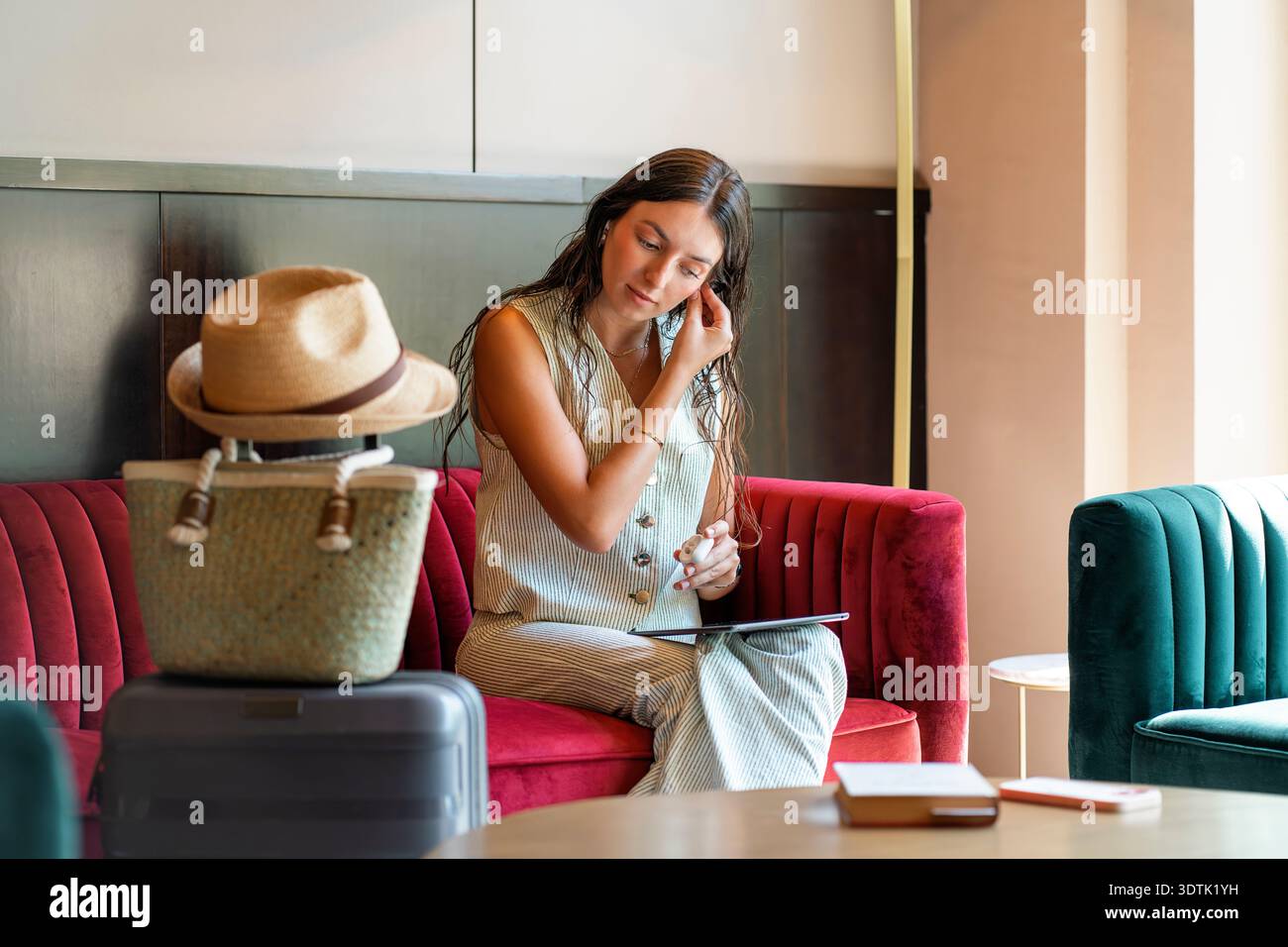 Una donna si siede comodamente sul divano di un hotel, inserendo gli auricolari mentre tiene in mano un tablet. Nelle vicinanze, una valigia e un cappello indicano il suo stile di vita di viaggio, combinin Foto Stock