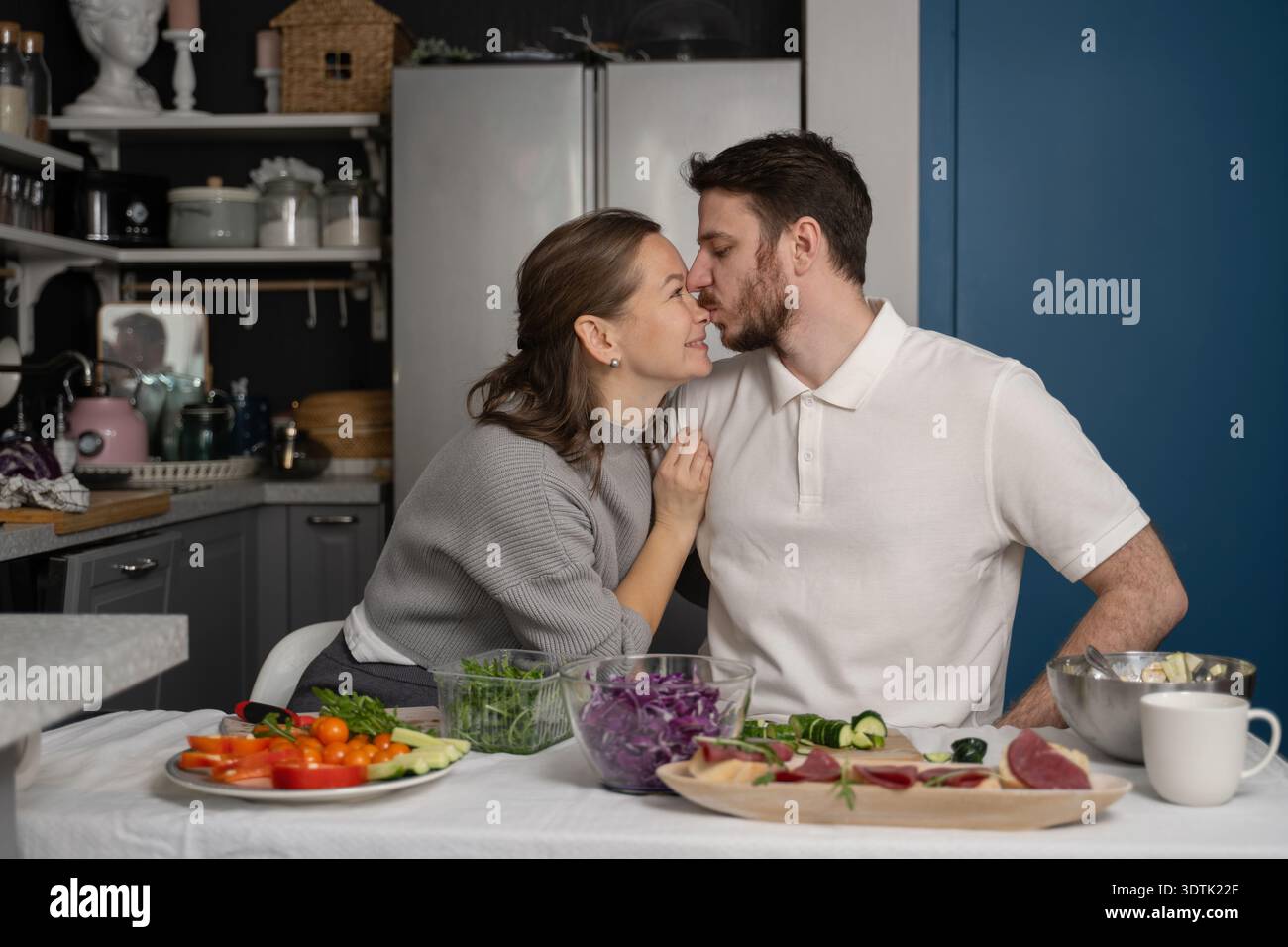 Una coppia amorevole condivide un bacio tenero nella loro accogliente cucina, circondata da ingredienti freschi e verdure vivaci. Il momento intimo cattura il calore Foto Stock