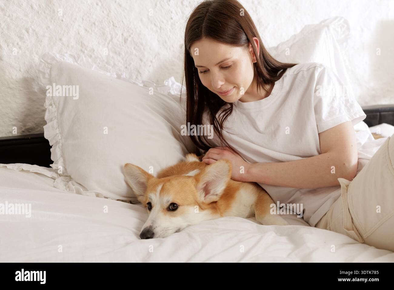 Giovane donna accarezzava il suo corgi riposando su morbidi letti in una camera da letto luminosa. Concetto di fiducia, comfort, cura degli animali e pacifica relazione tra animali umani e animali Foto Stock