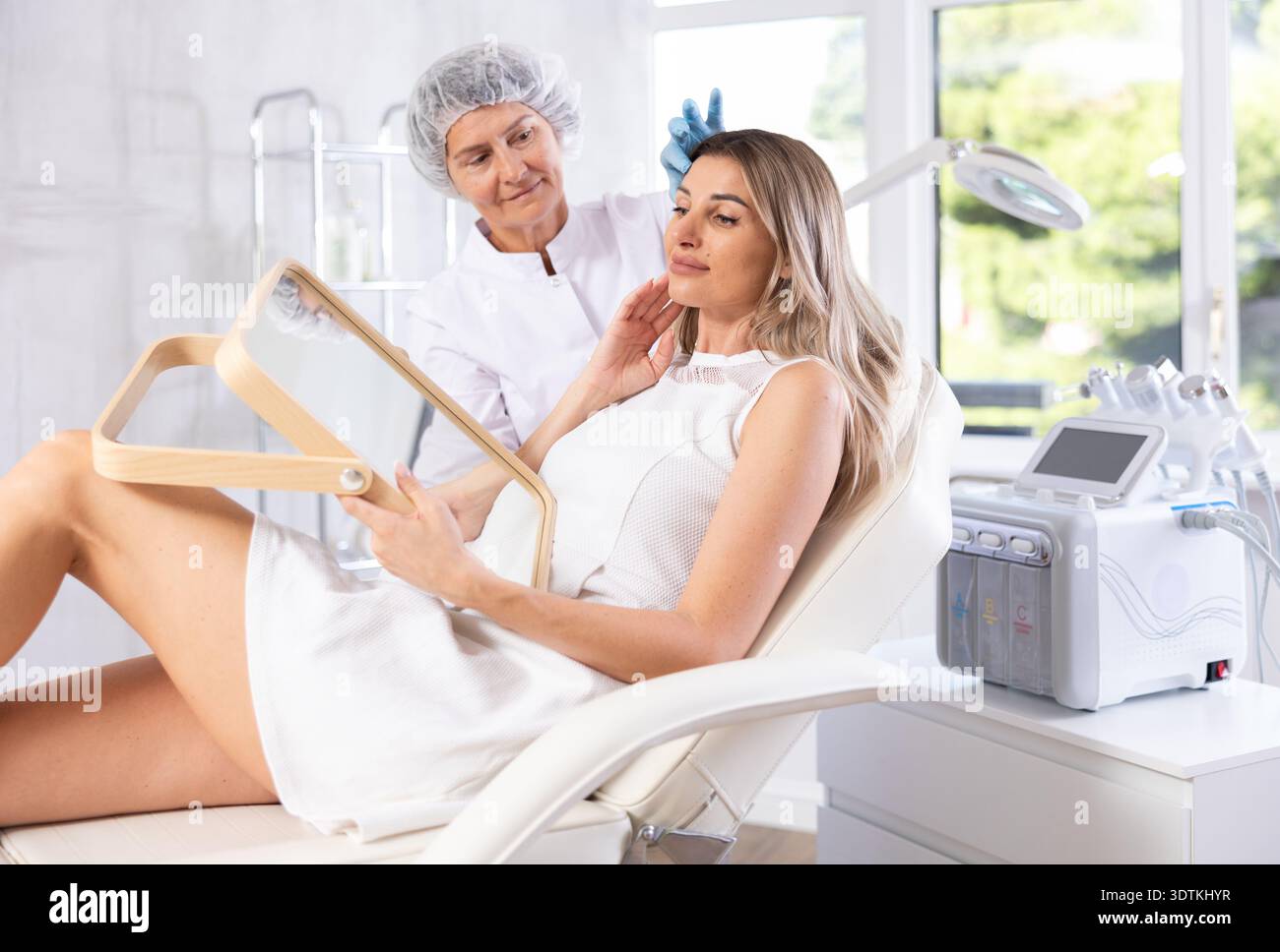 Giovane donna bionda soddisfatta che guarda nello specchio dopo un intervento di chirurgia plastica. Consultazione con chirurgo professionista Foto Stock