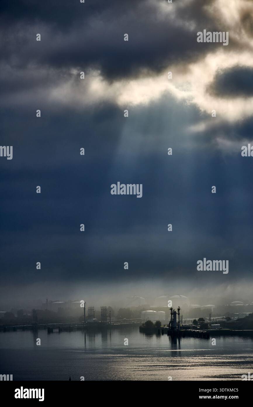 Una fotografia suggestiva e suggestiva che cattura pozzi di luce solare che penetrano attraverso nuvole scure e pesanti, illuminando un paesaggio industriale lontano Foto Stock